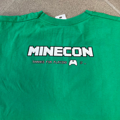 Minecraft 2015 Minecon London Green Tee is a green T-shirt made for Minecon 2015 in London, featuring Big Ben with a Creeper face on the front, with “Minecon London July 4-5” text in Minecraft-style lettering. The upper back displays “Minecon” with “Thanks for Playing” text below, while the sleeves carry a Mojang logo on one side and a Microsoft logo on the other.