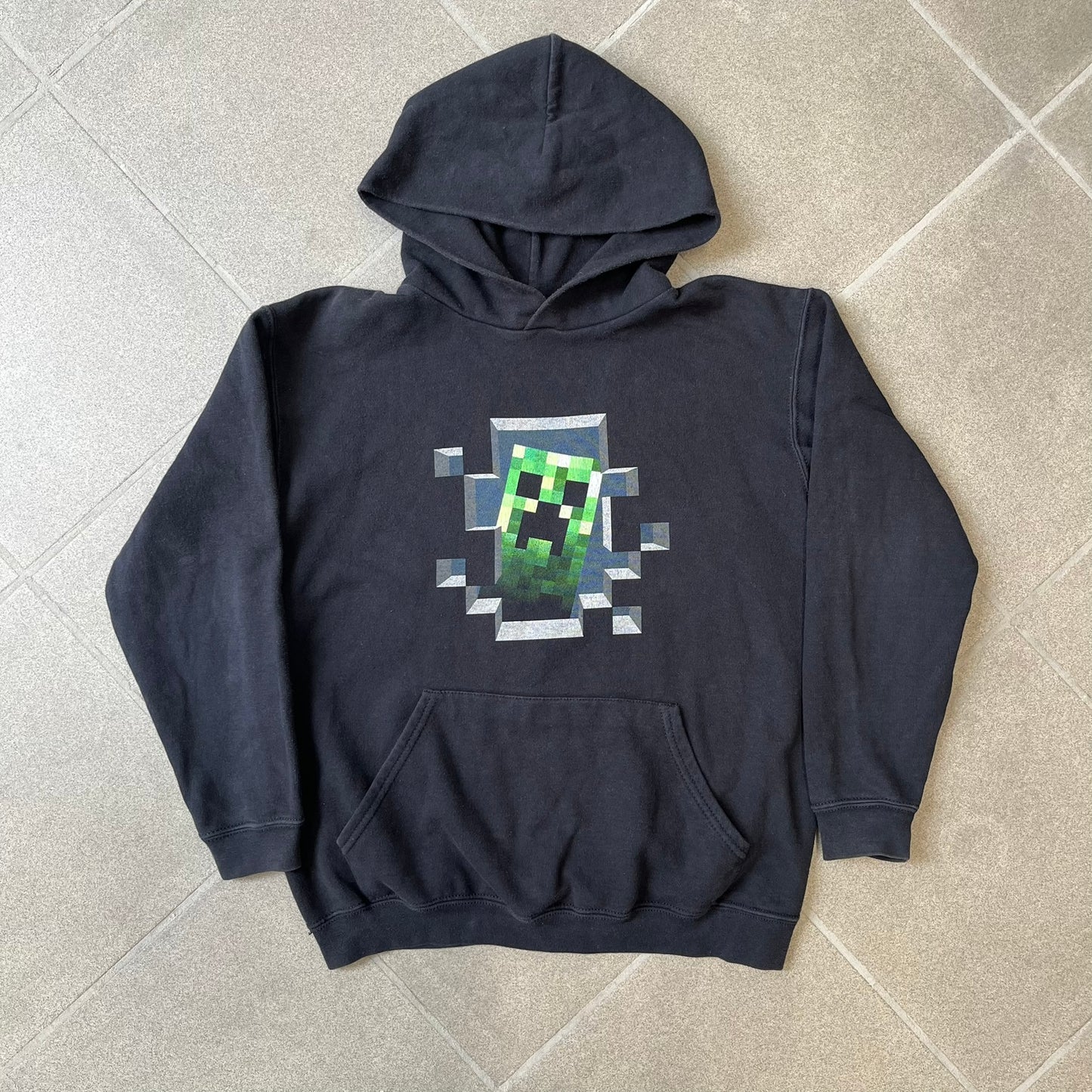 Minecraft Creeper 2013 J!NX Hoodie is a black 80/20 cotton-poly blend hoodie produced by Mojang and J!NX. The front features a Creeper graphic peeking through a pixelated hole designed to resemble in-game Minecraft blocks. This piece was released in 2013 as official Minecraft merchandise.