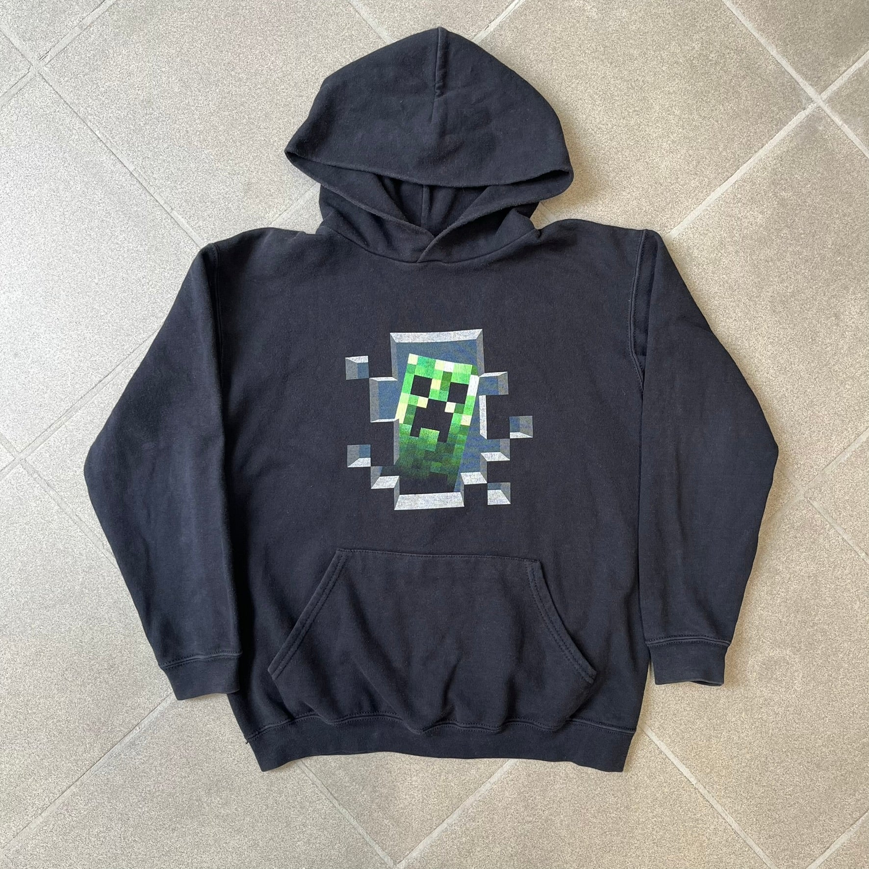 Minecraft Creeper 2013 J!NX Hoodie is a black 80/20 cotton-poly blend hoodie produced by Mojang and J!NX. The front features a Creeper graphic peeking through a pixelated hole designed to resemble in-game Minecraft blocks. This piece was released in 2013 as official Minecraft merchandise.