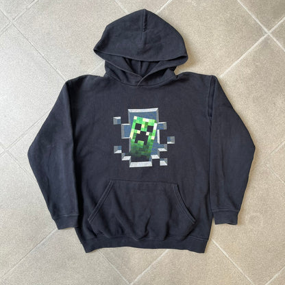 Minecraft Creeper 2013 J!NX Hoodie is a black 80/20 cotton-poly blend hoodie produced by Mojang and J!NX. The front features a Creeper graphic peeking through a pixelated hole designed to resemble in-game Minecraft blocks. This piece was released in 2013 as official Minecraft merchandise.