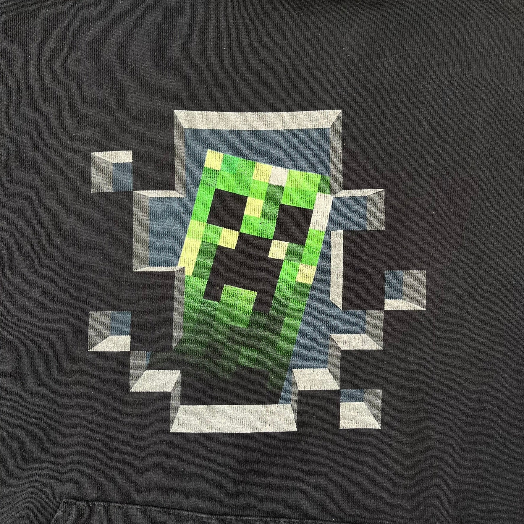 Minecraft Creeper 2013 J!NX Hoodie is a black 80/20 cotton-poly blend hoodie produced by Mojang and J!NX. The front features a Creeper graphic peeking through a pixelated hole designed to resemble in-game Minecraft blocks. This piece was released in 2013 as official Minecraft merchandise.