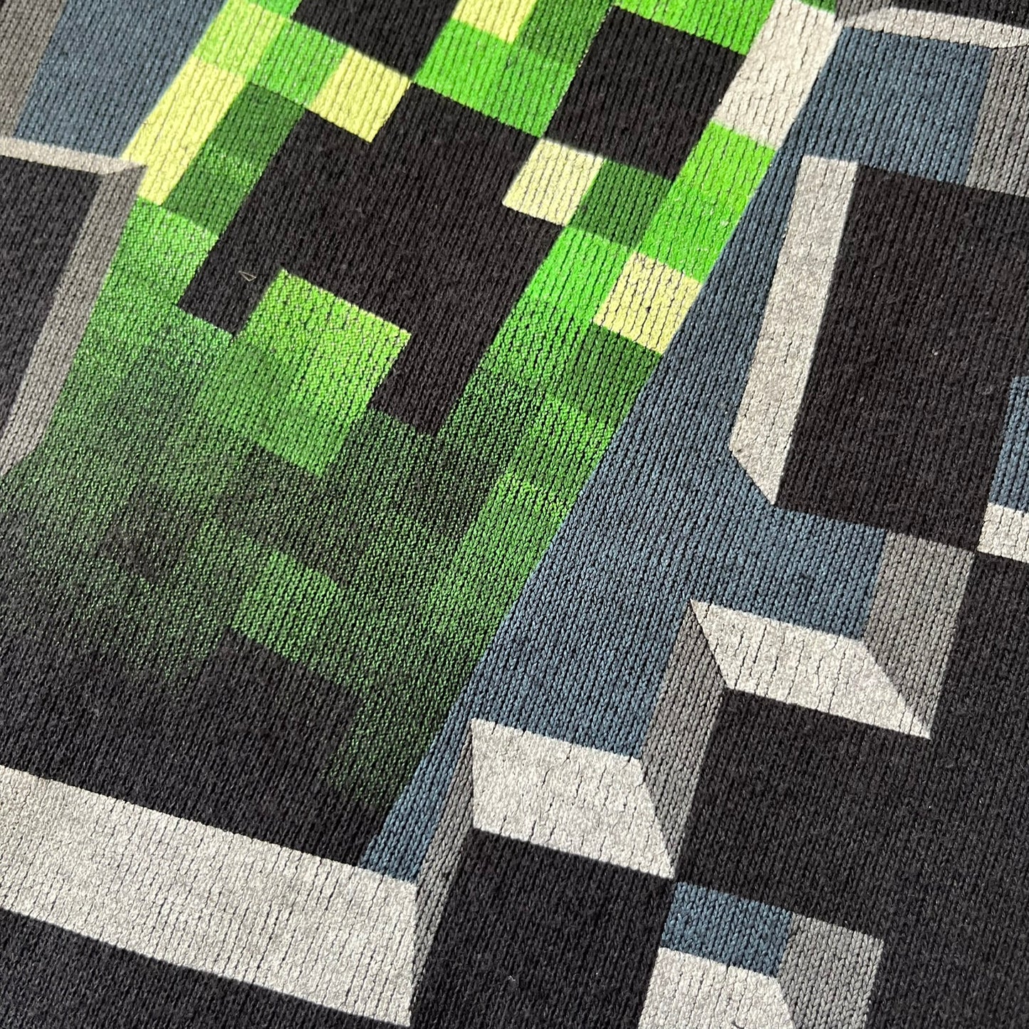 Minecraft Creeper 2013 J!NX Hoodie is a black 80/20 cotton-poly blend hoodie produced by Mojang and J!NX. The front features a Creeper graphic peeking through a pixelated hole designed to resemble in-game Minecraft blocks. This piece was released in 2013 as official Minecraft merchandise.