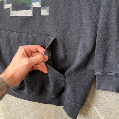 Minecraft Creeper 2013 J!NX Hoodie is a black 80/20 cotton-poly blend hoodie produced by Mojang and J!NX. The front features a Creeper graphic peeking through a pixelated hole designed to resemble in-game Minecraft blocks. This piece was released in 2013 as official Minecraft merchandise.