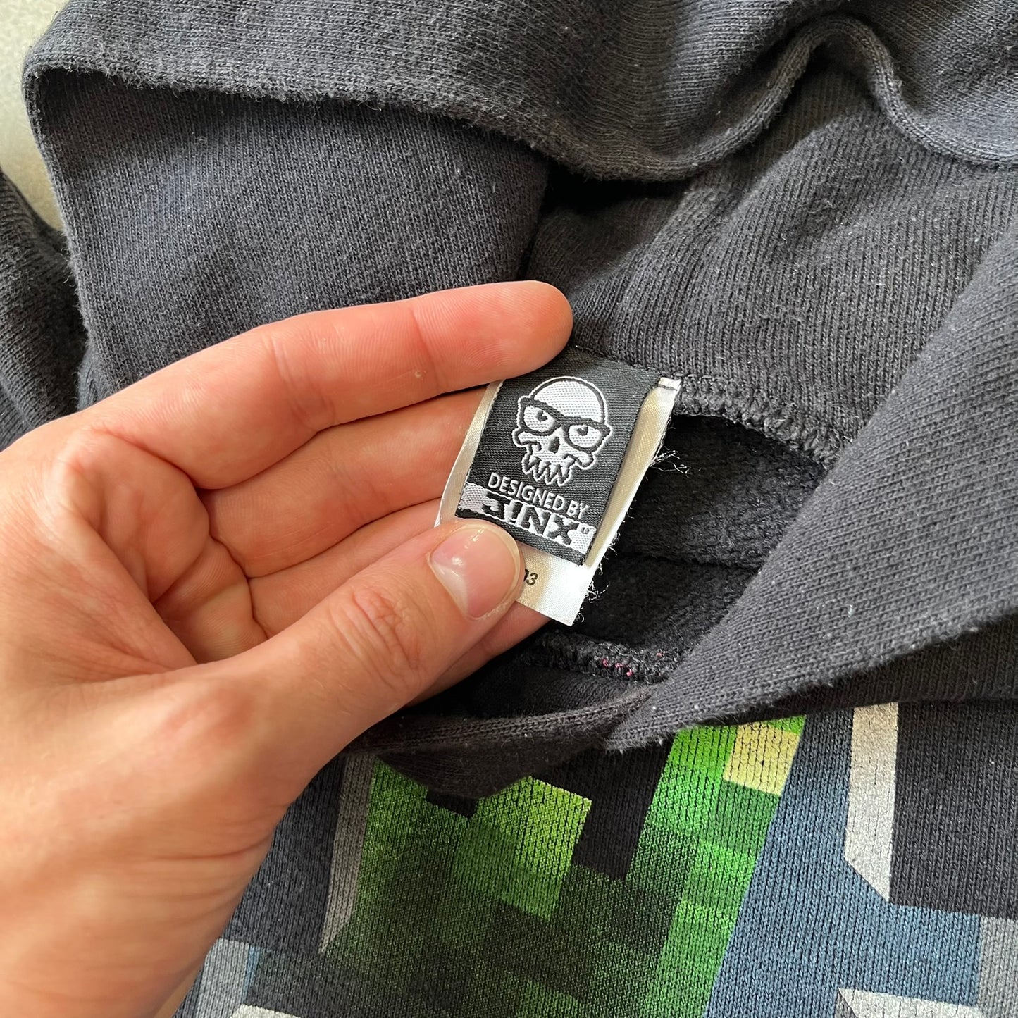 Minecraft Creeper 2013 J!NX Hoodie is a black 80/20 cotton-poly blend hoodie produced by Mojang and J!NX. The front features a Creeper graphic peeking through a pixelated hole designed to resemble in-game Minecraft blocks. This piece was released in 2013 as official Minecraft merchandise.