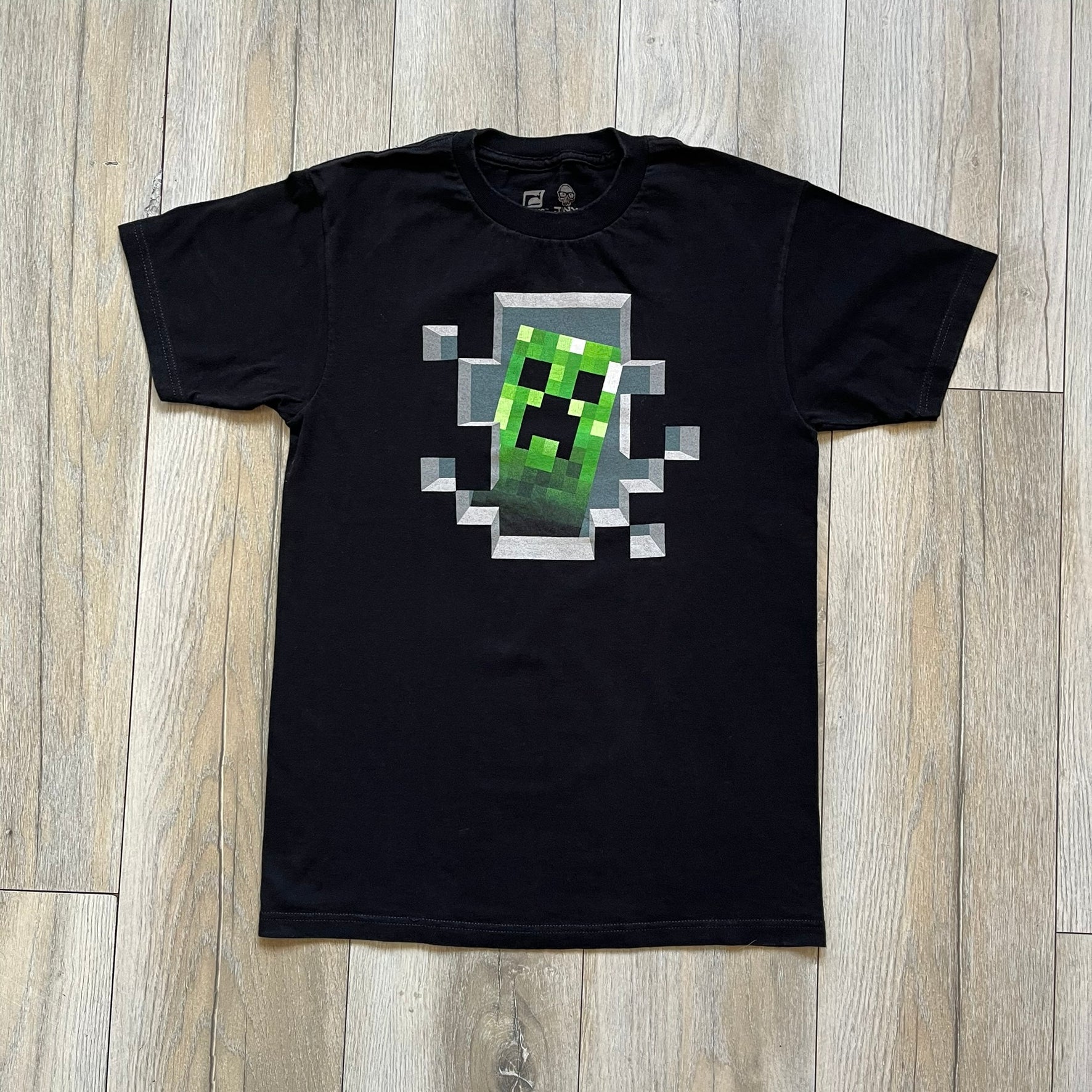 Minecraft Creeper 2013 J!NX Tee is a black 100% cotton T-shirt produced by Mojang and J!NX. The front features a Creeper graphic peeking through a pixelated hole designed to resemble in-game Minecraft blocks. This piece was released in 2013 as official Minecraft merchandise.