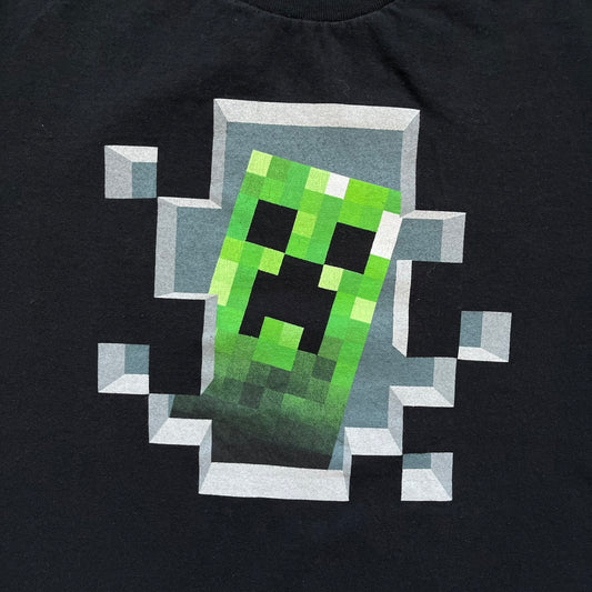 Minecraft Creeper 2013 J!NX Tee is a black 100% cotton T-shirt produced by Mojang and J!NX. The front features a Creeper graphic peeking through a pixelated hole designed to resemble in-game Minecraft blocks. This piece was released in 2013 as official Minecraft merchandise.