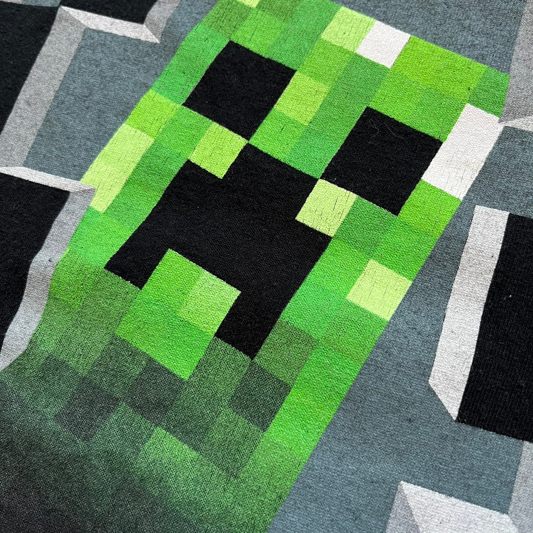 Minecraft Creeper 2013 J!NX Tee is a black 100% cotton T-shirt produced by Mojang and J!NX. The front features a Creeper graphic peeking through a pixelated hole designed to resemble in-game Minecraft blocks. This piece was released in 2013 as official Minecraft merchandise.