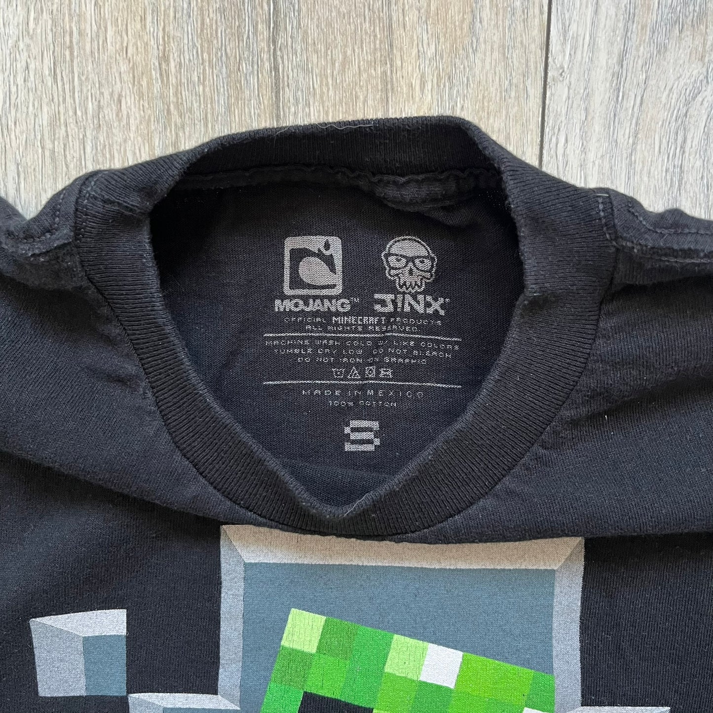 Minecraft Creeper 2013 J!NX Tee is a black 100% cotton T-shirt produced by Mojang and J!NX. The front features a Creeper graphic peeking through a pixelated hole designed to resemble in-game Minecraft blocks. This piece was released in 2013 as official Minecraft merchandise.