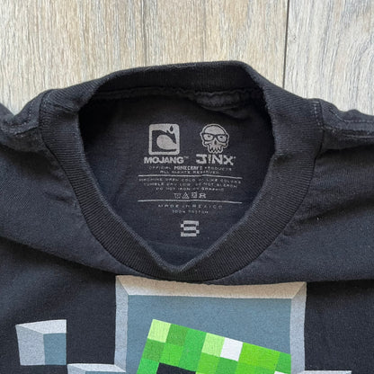Minecraft Creeper 2013 J!NX Tee is a black 100% cotton T-shirt produced by Mojang and J!NX. The front features a Creeper graphic peeking through a pixelated hole designed to resemble in-game Minecraft blocks. This piece was released in 2013 as official Minecraft merchandise.