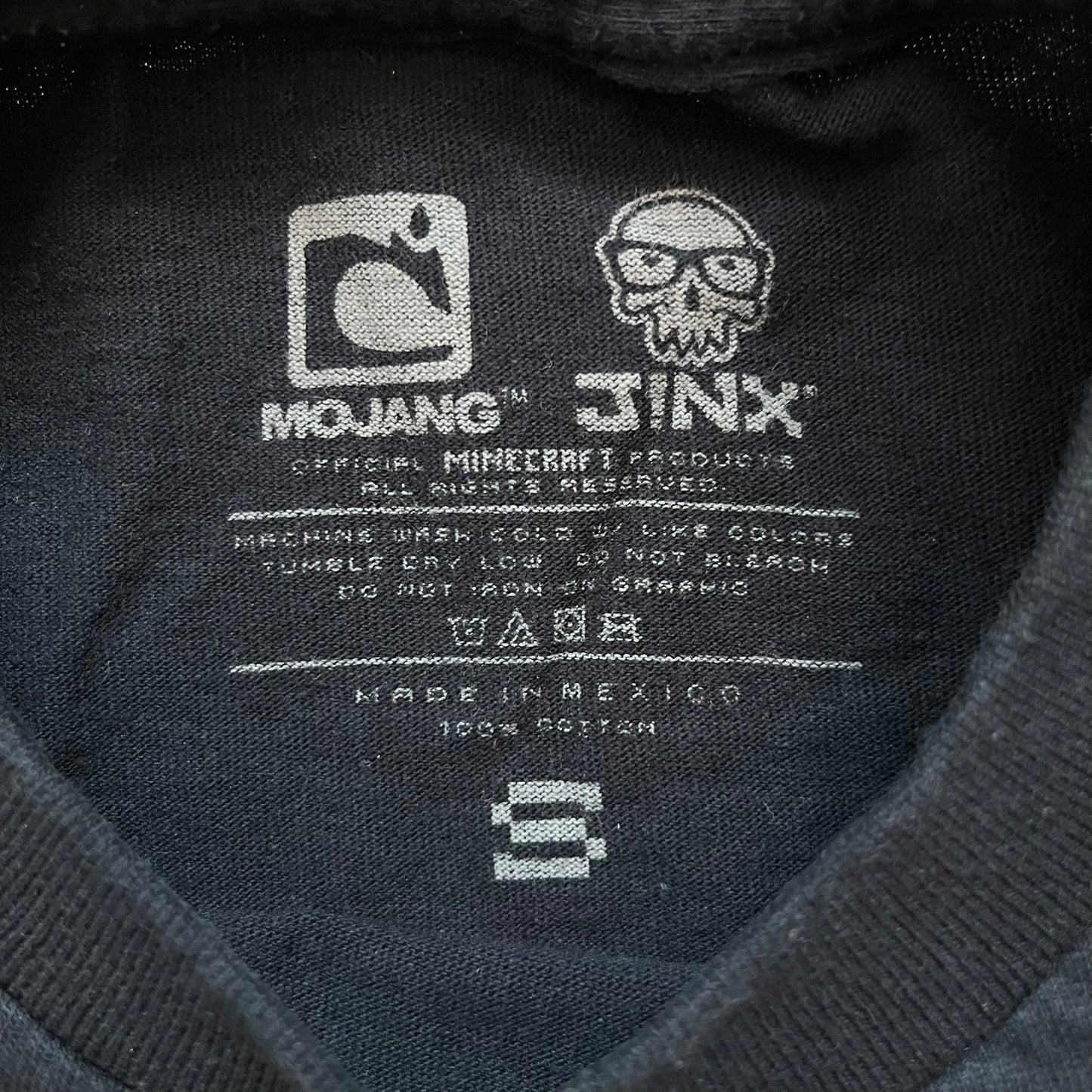 Minecraft Creeper 2013 J!NX Tee is a black 100% cotton T-shirt produced by Mojang and J!NX. The front features a Creeper graphic peeking through a pixelated hole designed to resemble in-game Minecraft blocks. This piece was released in 2013 as official Minecraft merchandise.