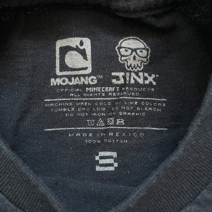 Minecraft Creeper 2013 J!NX Tee is a black 100% cotton T-shirt produced by Mojang and J!NX. The front features a Creeper graphic peeking through a pixelated hole designed to resemble in-game Minecraft blocks. This piece was released in 2013 as official Minecraft merchandise.