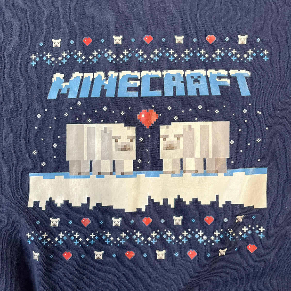 Minecraft Polar Bears Navy Crewneck features a navy construction made on a Fruit of the Loom blank. The front has a pattern of hearts and polar bear heads, with an icy Minecraft logo topped with snow and two polar bears standing on snow with a heart between them. This is not an official Minecraft piece.
