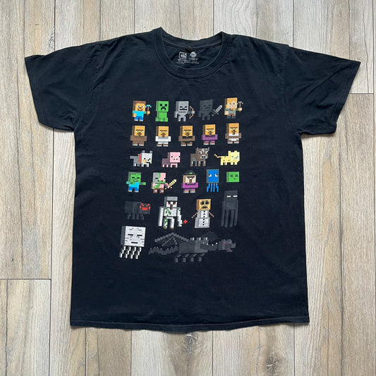 Minecraft Sprites 2016 J!NX Tee is a black cotton T-shirt with a slightly washed finish. The front features a large rectangular grid-style print made up of multiple Minecraft character sprites arranged in a structured pattern. It includes Steve, villagers, hostile mobs, and animals in a simplified pixel-art style.
