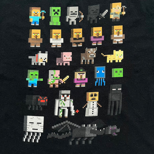 Minecraft Sprites 2016 J!NX Tee is a black cotton T-shirt with a slightly washed finish. The front features a large rectangular grid-style print made up of multiple Minecraft character sprites arranged in a structured pattern. It includes Steve, villagers, hostile mobs, and animals in a simplified pixel-art style.
