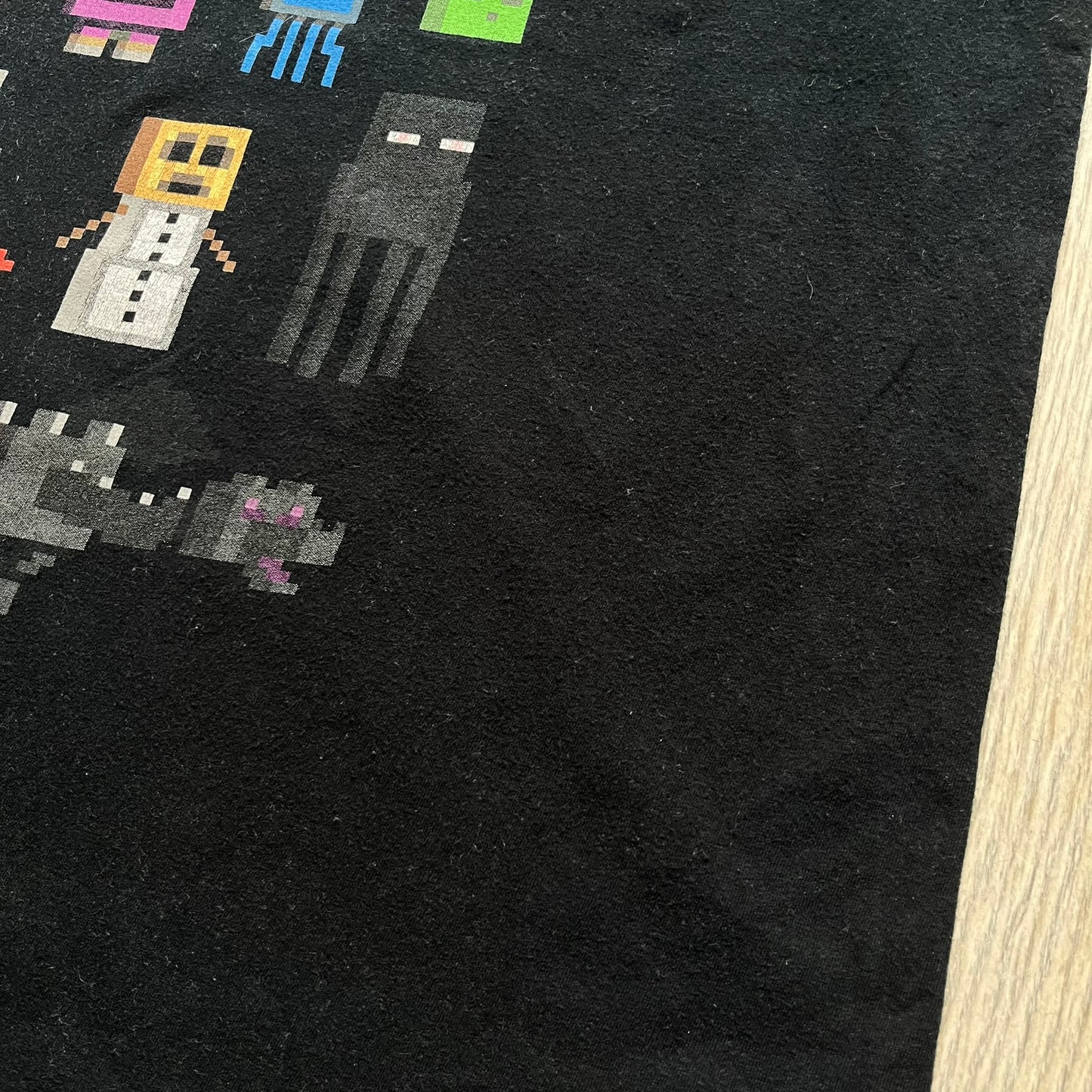 Minecraft Sprites 2016 J!NX Tee is a black cotton T-shirt with a slightly washed finish. The front features a large rectangular grid-style print made up of multiple Minecraft character sprites arranged in a structured pattern. It includes Steve, villagers, hostile mobs, and animals in a simplified pixel-art style.
