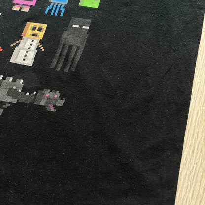 Minecraft Sprites 2016 J!NX Tee is a black cotton T-shirt with a slightly washed finish. The front features a large rectangular grid-style print made up of multiple Minecraft character sprites arranged in a structured pattern. It includes Steve, villagers, hostile mobs, and animals in a simplified pixel-art style.
