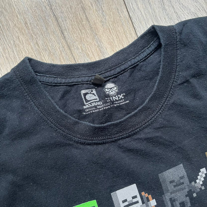 Minecraft Sprites 2016 J!NX Tee is a black cotton T-shirt with a slightly washed finish. The front features a large rectangular grid-style print made up of multiple Minecraft character sprites arranged in a structured pattern. It includes Steve, villagers, hostile mobs, and animals in a simplified pixel-art style.
