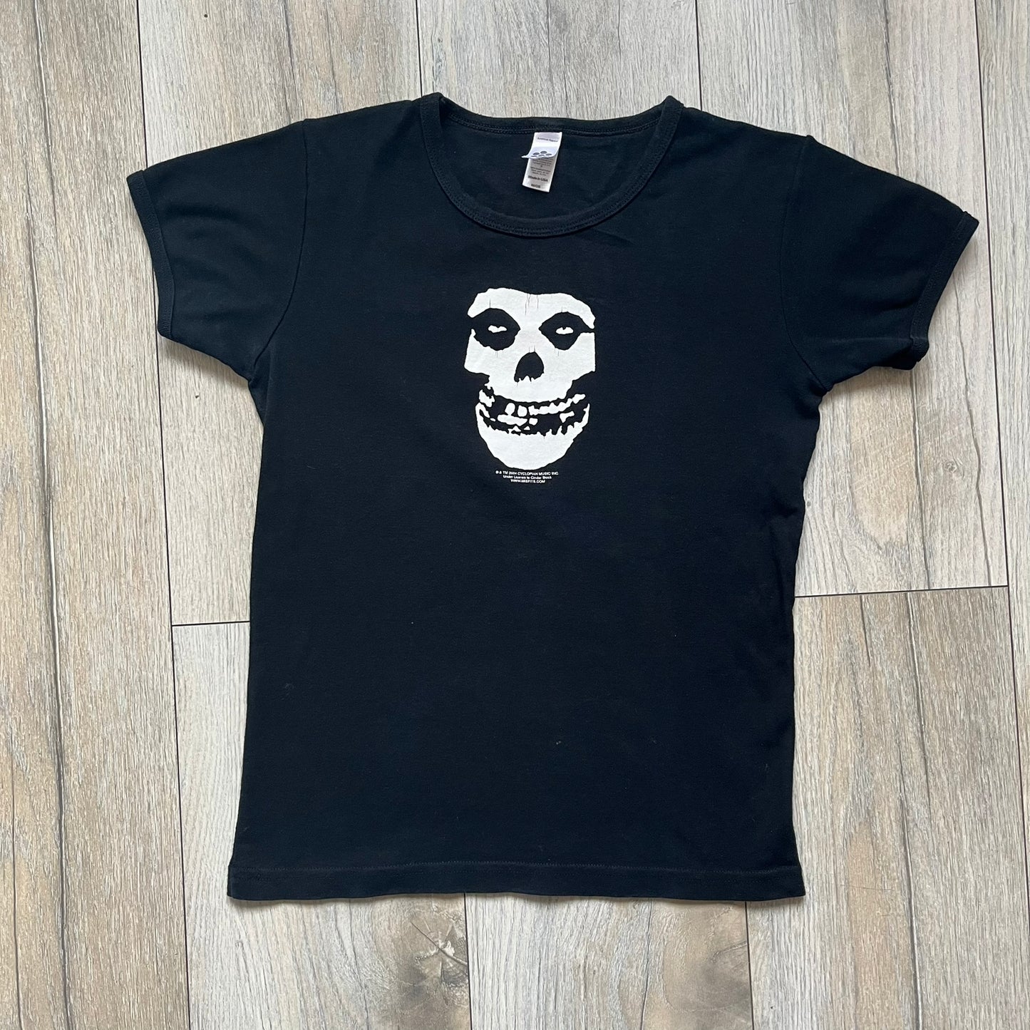 Misfits 2004 Fiend Skull Black Tee is a black T-shirt made in the USA on an American Apparel Classic Girl blank. The front features the band’s white Fiend Skull graphic printed on the chest. The shirt includes 2004 Cyclopian Music Inc. licensing and a Misfits neck tag.