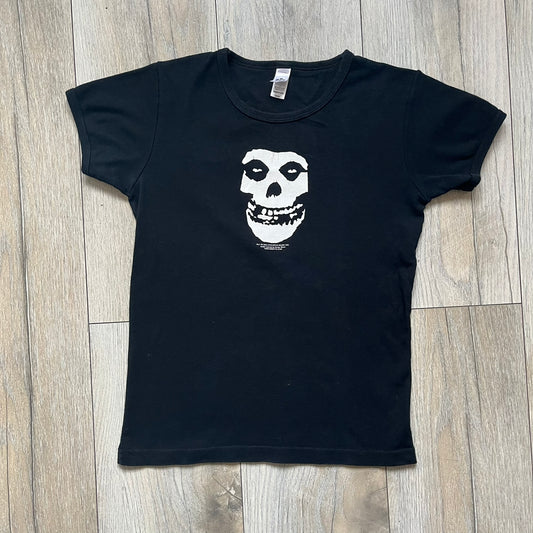 Misfits 2004 Fiend Skull Black Tee is a black T-shirt made in the USA on an American Apparel Classic Girl blank. The front features the band’s white Fiend Skull graphic printed on the chest. The shirt includes 2004 Cyclopian Music Inc. licensing and a Misfits neck tag.