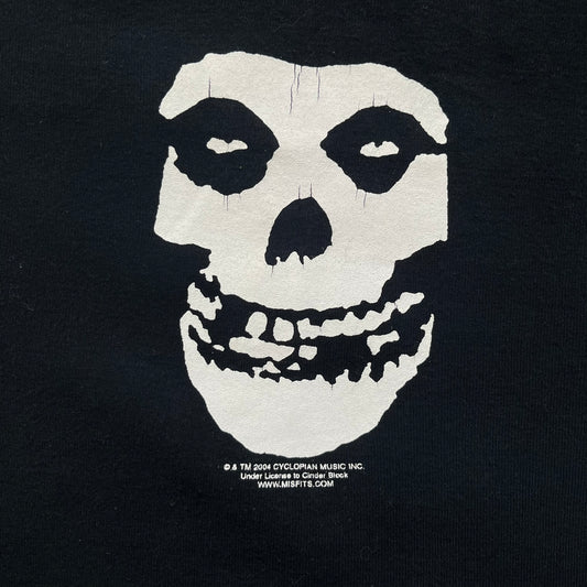 Misfits 2004 Fiend Skull Black Tee is a black T-shirt made in the USA on an American Apparel Classic Girl blank. The front features the band’s white Fiend Skull graphic printed on the chest. The shirt includes 2004 Cyclopian Music Inc. licensing and a Misfits neck tag.