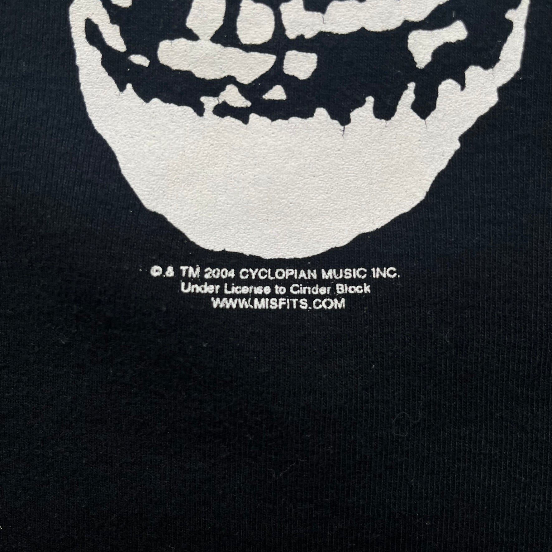 Misfits 2004 Fiend Skull Black Tee is a black T-shirt made in the USA on an American Apparel Classic Girl blank. The front features the band’s white Fiend Skull graphic printed on the chest. The shirt includes 2004 Cyclopian Music Inc. licensing and a Misfits neck tag.