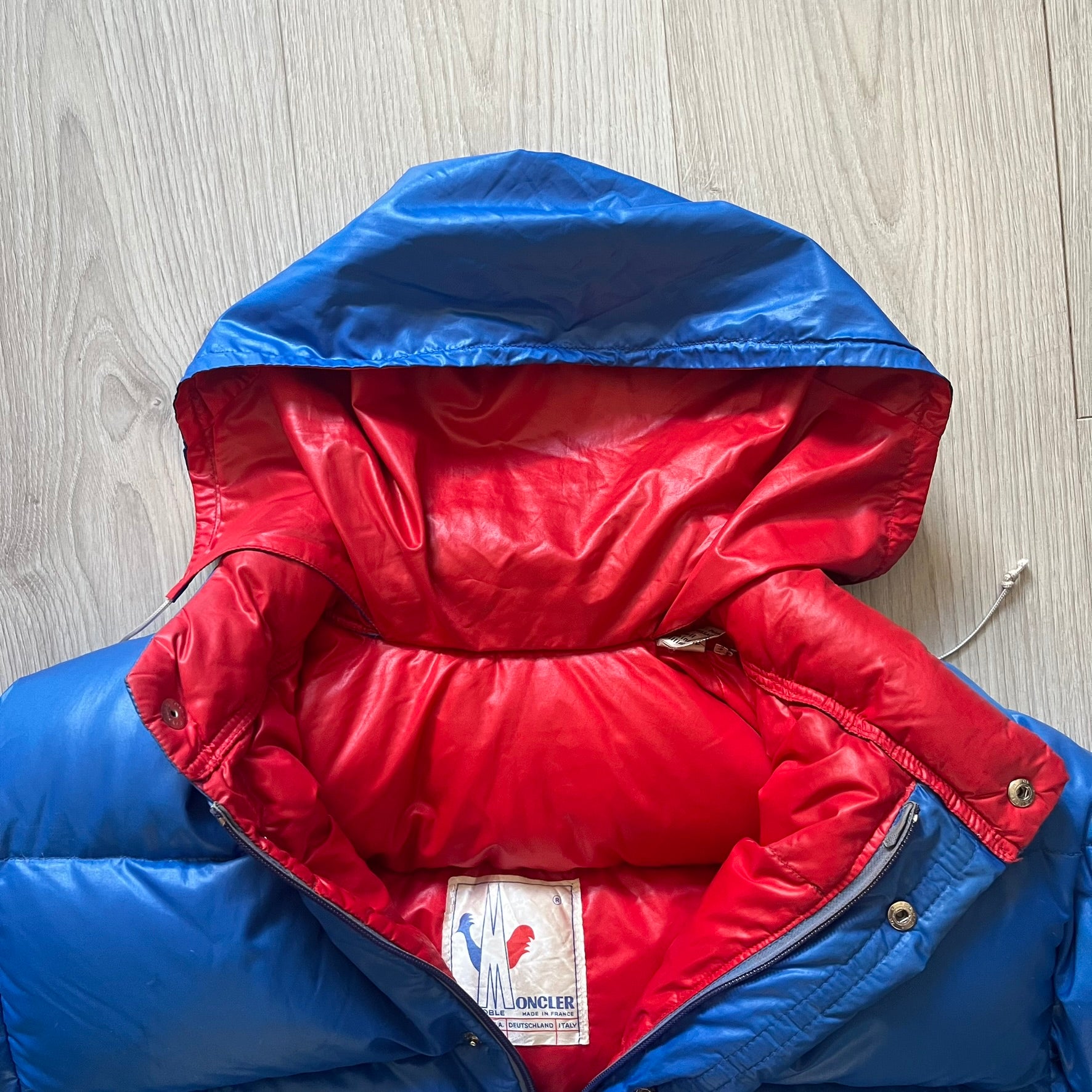 Vintage 1980s Moncler Grenoble down jacket in blue with a contrasting red lining. Features a foldable thin hood hidden in the back, silver-tone buttons and zippers, and the original Moncler patch on the sleeve. A rare French-made piece with authentic Grenoble labels, distinct from the brand’s modern ski line.