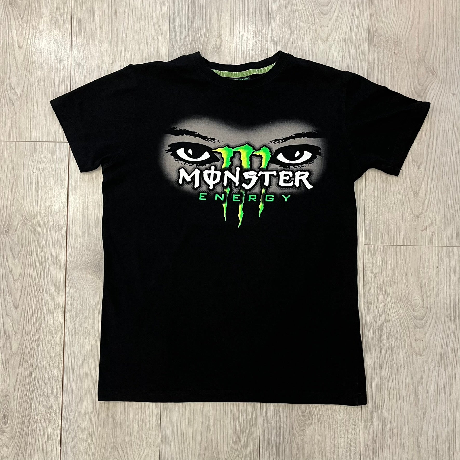 Monster Energy Eyes Tee in black, featuring a graphic of a woman’s eyes with the Monster logo and text on the front in gray and white tones. Green shoulder stitching and a Monster Energy logo on the back under the collar. Made from 95% cotton and 5% Lycra.