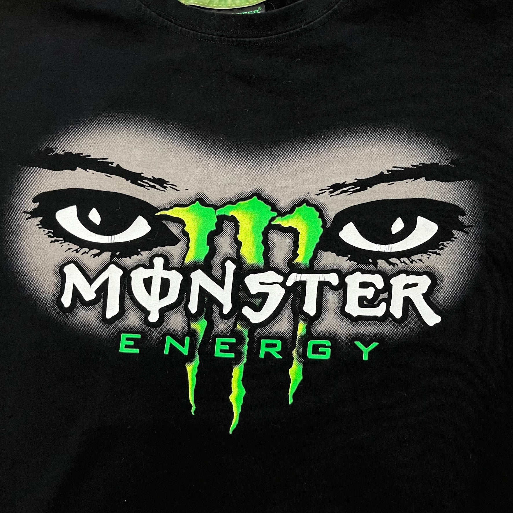 Monster Energy Eyes Tee in black, featuring a graphic of a woman’s eyes with the Monster logo and text on the front in gray and white tones. Green shoulder stitching and a Monster Energy logo on the back under the collar. Made from 95% cotton and 5% Lycra.