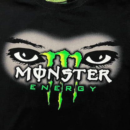 Monster Energy Eyes Tee in black, featuring a graphic of a woman’s eyes with the Monster logo and text on the front in gray and white tones. Green shoulder stitching and a Monster Energy logo on the back under the collar. Made from 95% cotton and 5% Lycra.
