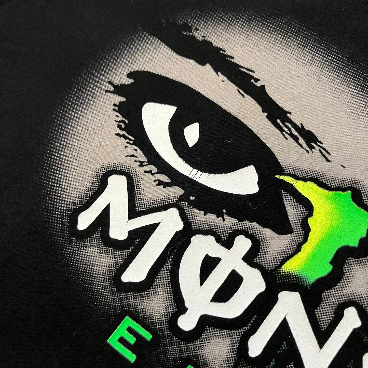 Monster Energy Eyes Tee in black, featuring a graphic of a woman’s eyes with the Monster logo and text on the front in gray and white tones. Green shoulder stitching and a Monster Energy logo on the back under the collar. Made from 95% cotton and 5% Lycra.