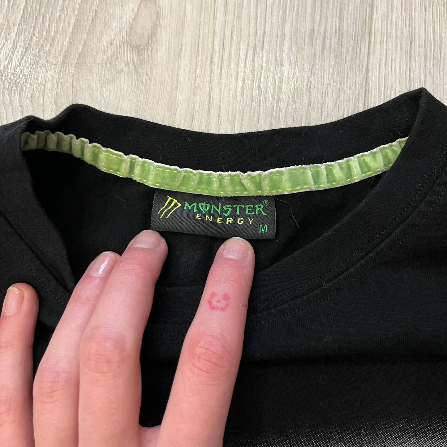 Monster Energy Eyes Tee in black, featuring a graphic of a woman’s eyes with the Monster logo and text on the front in gray and white tones. Green shoulder stitching and a Monster Energy logo on the back under the collar. Made from 95% cotton and 5% Lycra.