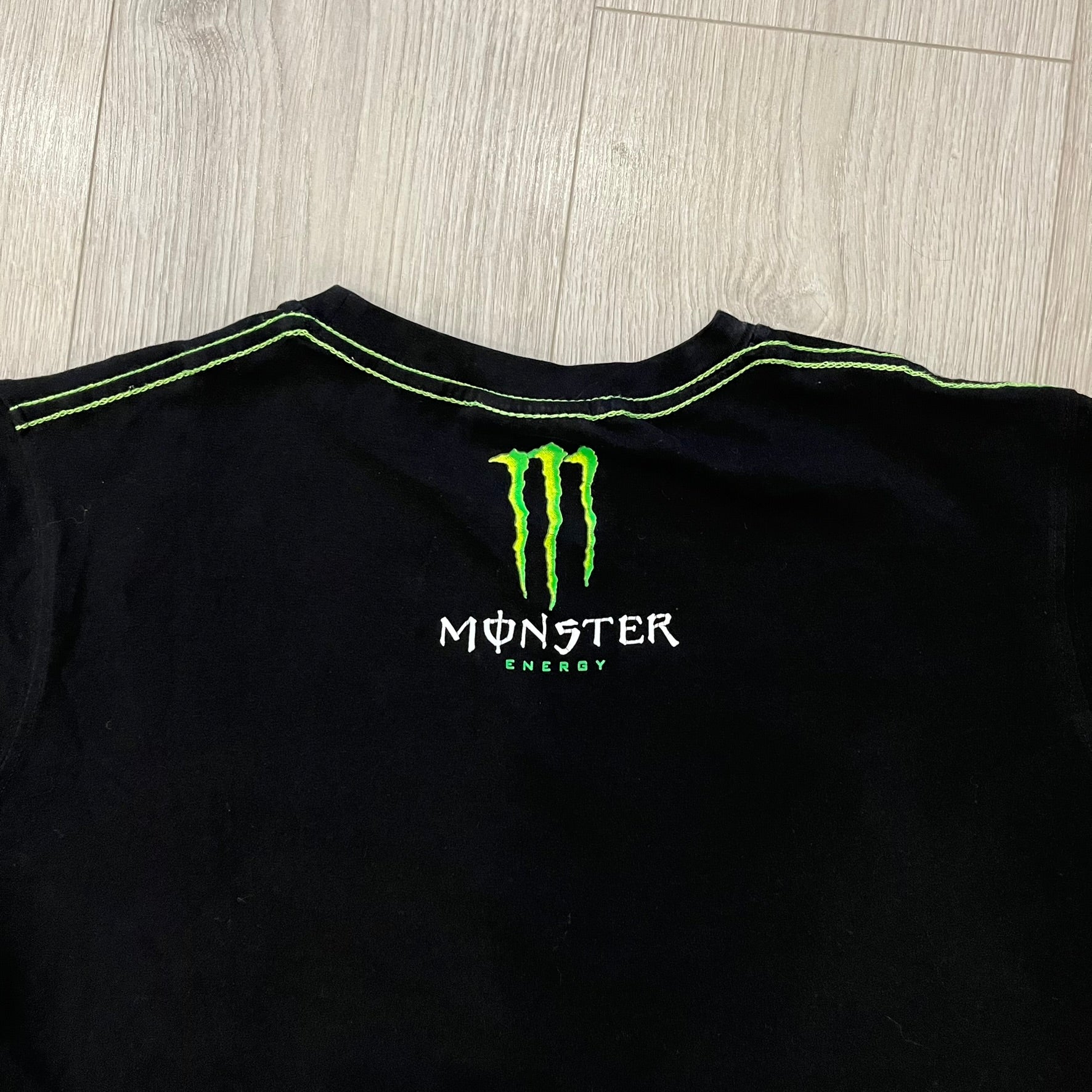 Monster Energy Eyes Tee in black, featuring a graphic of a woman’s eyes with the Monster logo and text on the front in gray and white tones. Green shoulder stitching and a Monster Energy logo on the back under the collar. Made from 95% cotton and 5% Lycra.