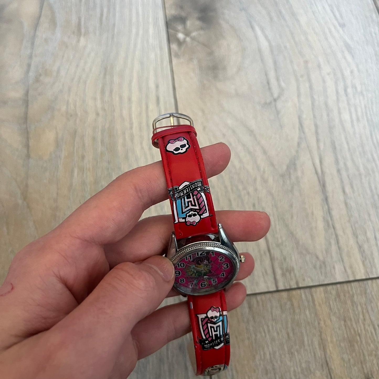 Monster High Red & Pink Kids Watch is a children’s wristwatch featuring a red strap printed with Monster High logos and character graphics. The watch has a silver-tone case with a pink dial, black numerals, and white hands, along with a Monster High character graphic and logo on the face. It comes packaged in a pink box with full Monster High branding.
