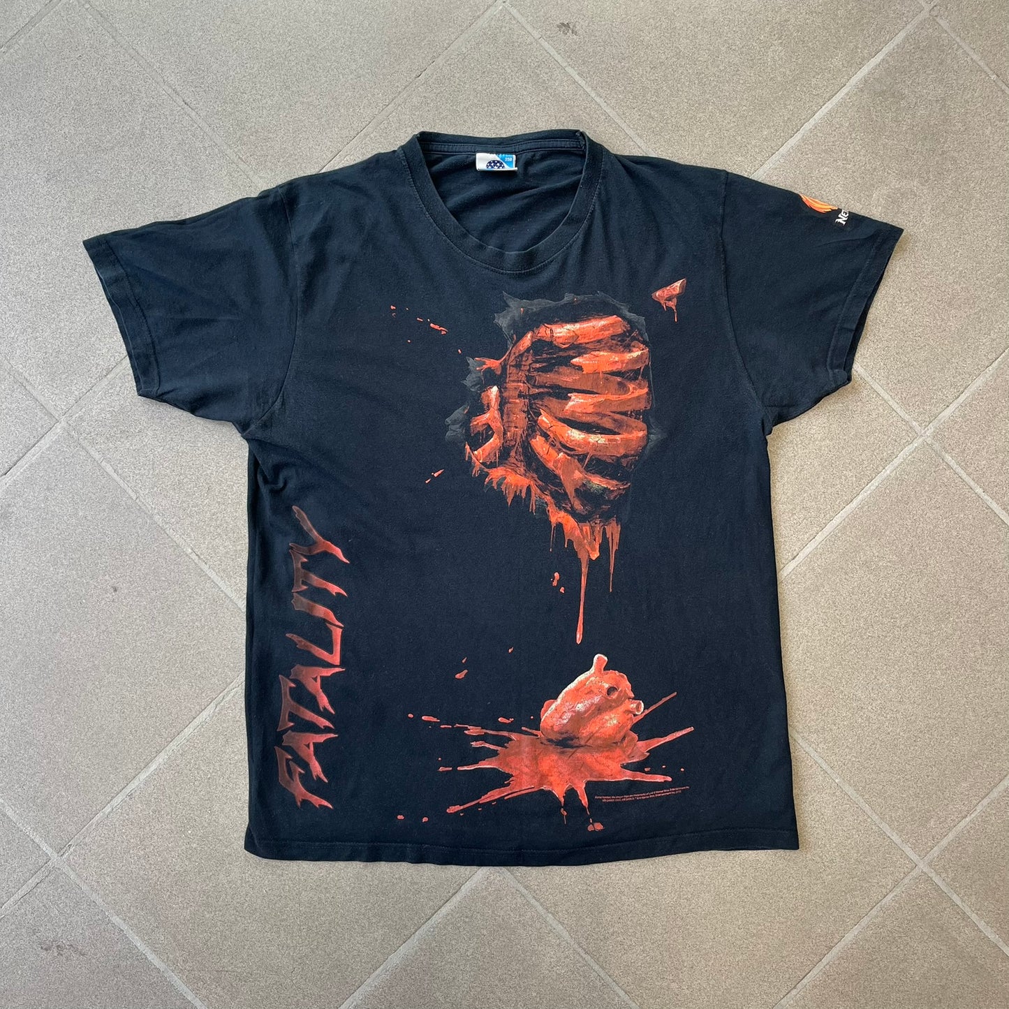Mortal Kombat 9 Fatality Truck Tour 2011 Tee features a black T-shirt with a front graphic of a broken red ribcage and heart on a blood-covered surface. A vertical “Fatality” graphic is printed along the side, with Warner Bros. licensing text underneath the heart. One sleeve features the NetherRealm Studios logo. The back has the classic “Finish Him” text, Mortal Kombat logo, and matching licensing details.
