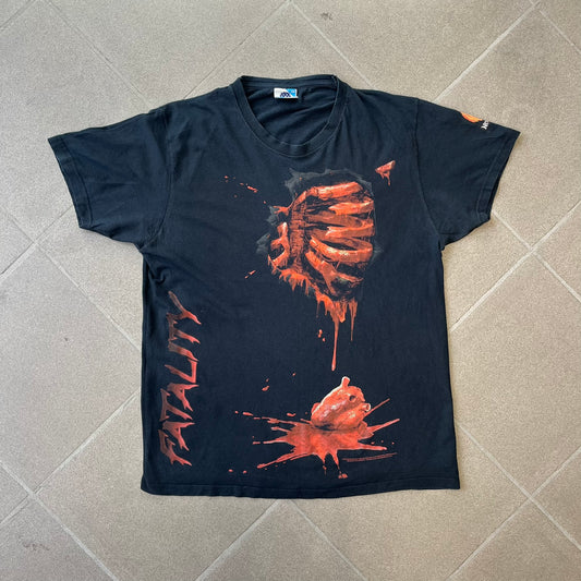 Mortal Kombat 9 Fatality Truck Tour 2011 Tee features a black T-shirt with a front graphic of a broken red ribcage and heart on a blood-covered surface. A vertical “Fatality” graphic is printed along the side, with Warner Bros. licensing text underneath the heart. One sleeve features the NetherRealm Studios logo. The back has the classic “Finish Him” text, Mortal Kombat logo, and matching licensing details.
