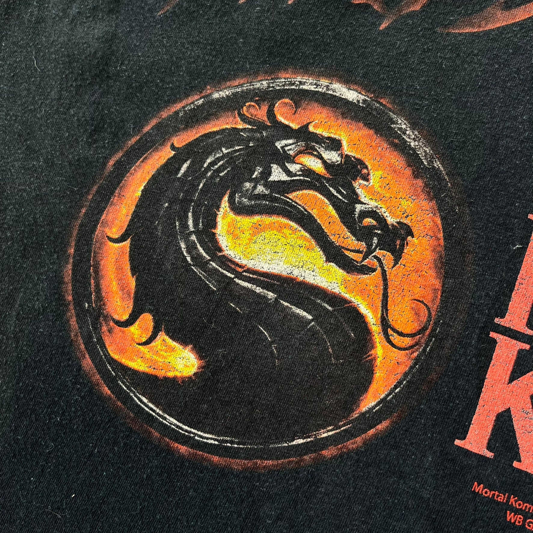 Mortal Kombat 9 Fatality Truck Tour 2011 Tee features a black T-shirt with a front graphic of a broken red ribcage and heart on a blood-covered surface. A vertical “Fatality” graphic is printed along the side, with Warner Bros. licensing text underneath the heart. One sleeve features the NetherRealm Studios logo. The back has the classic “Finish Him” text, Mortal Kombat logo, and matching licensing details.
