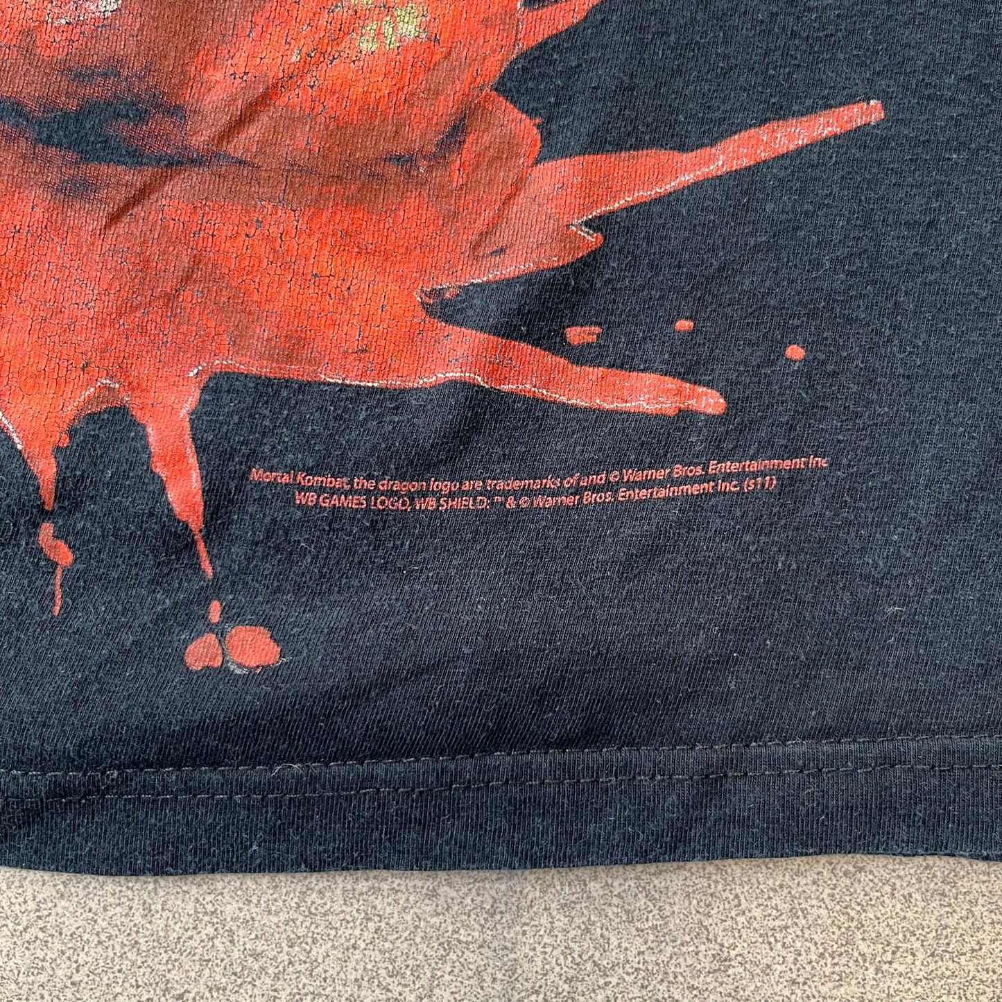 Mortal Kombat 9 Fatality Truck Tour 2011 Tee features a black T-shirt with a front graphic of a broken red ribcage and heart on a blood-covered surface. A vertical “Fatality” graphic is printed along the side, with Warner Bros. licensing text underneath the heart. One sleeve features the NetherRealm Studios logo. The back has the classic “Finish Him” text, Mortal Kombat logo, and matching licensing details.

