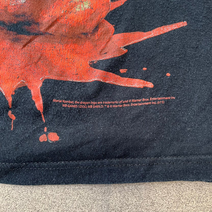 Mortal Kombat 9 Fatality Truck Tour 2011 Tee features a black T-shirt with a front graphic of a broken red ribcage and heart on a blood-covered surface. A vertical “Fatality” graphic is printed along the side, with Warner Bros. licensing text underneath the heart. One sleeve features the NetherRealm Studios logo. The back has the classic “Finish Him” text, Mortal Kombat logo, and matching licensing details.
