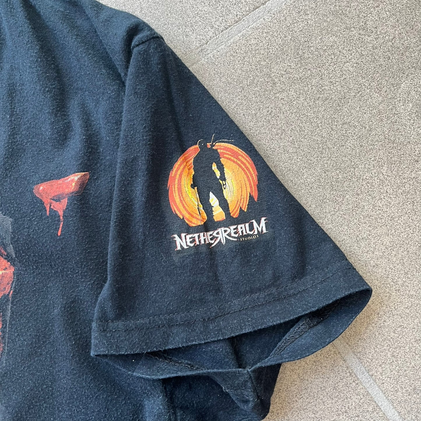 Mortal Kombat 9 Fatality Truck Tour 2011 Tee features a black T-shirt with a front graphic of a broken red ribcage and heart on a blood-covered surface. A vertical “Fatality” graphic is printed along the side, with Warner Bros. licensing text underneath the heart. One sleeve features the NetherRealm Studios logo. The back has the classic “Finish Him” text, Mortal Kombat logo, and matching licensing details.
