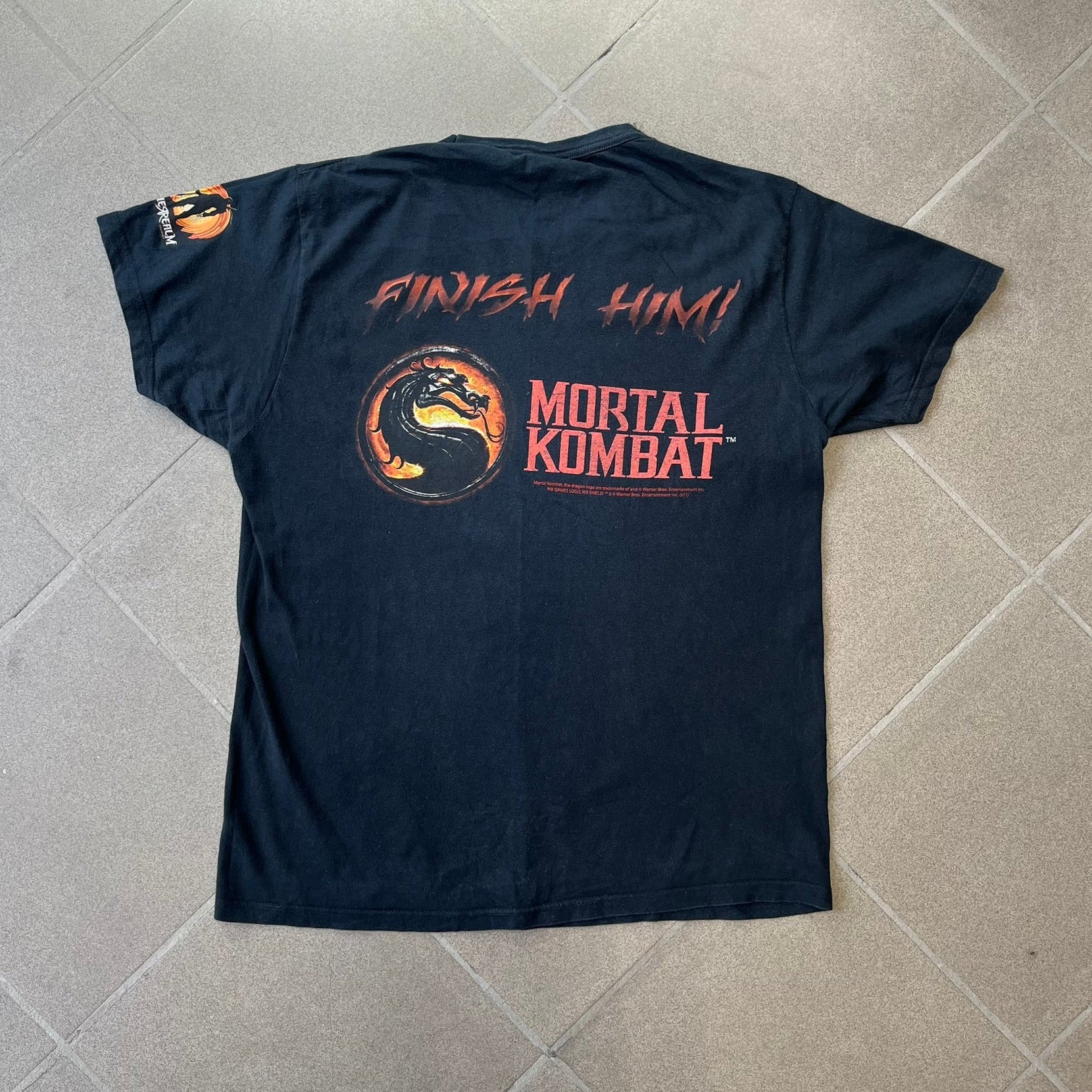 Mortal Kombat 9 Fatality Truck Tour 2011 Tee features a black T-shirt with a front graphic of a broken red ribcage and heart on a blood-covered surface. A vertical “Fatality” graphic is printed along the side, with Warner Bros. licensing text underneath the heart. One sleeve features the NetherRealm Studios logo. The back has the classic “Finish Him” text, Mortal Kombat logo, and matching licensing details.
