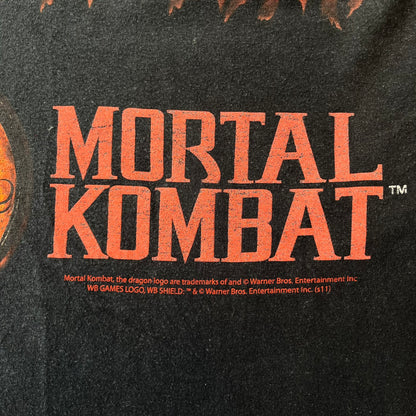 Mortal Kombat 9 Fatality Truck Tour 2011 Tee features a black T-shirt with a front graphic of a broken red ribcage and heart on a blood-covered surface. A vertical “Fatality” graphic is printed along the side, with Warner Bros. licensing text underneath the heart. One sleeve features the NetherRealm Studios logo. The back has the classic “Finish Him” text, Mortal Kombat logo, and matching licensing details.
