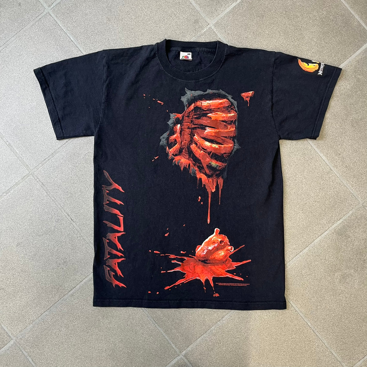 Mortal Kombat 9 Fatality Truck Tour 2011 Tee features a black T-shirt with a front graphic of a broken red ribcage and heart on a blood-covered surface. A vertical “Fatality” graphic is printed along the side, with Warner Bros. licensing text underneath the heart. One sleeve features the NetherRealm Studios logo. The back has the classic “Finish Him” text, Mortal Kombat logo, and matching licensing details.
