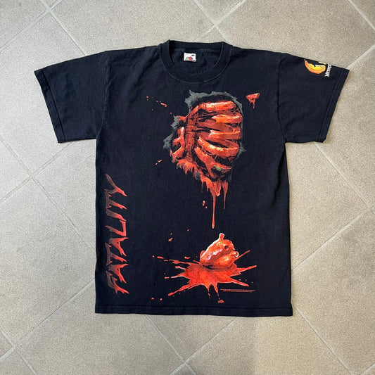 Mortal Kombat 9 Fatality Truck Tour 2011 Tee features a black T-shirt with a front graphic of a broken red ribcage and heart on a blood-covered surface. A vertical “Fatality” graphic is printed along the side, with Warner Bros. licensing text underneath the heart. One sleeve features the NetherRealm Studios logo. The back has the classic “Finish Him” text, Mortal Kombat logo, and matching licensing details.
