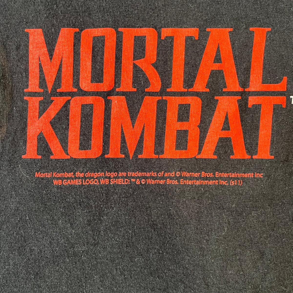 Mortal Kombat 9 Fatality Truck Tour 2011 Tee features a black T-shirt with a front graphic of a broken red ribcage and heart on a blood-covered surface. A vertical “Fatality” graphic is printed along the side, with Warner Bros. licensing text underneath the heart. One sleeve features the NetherRealm Studios logo. The back has the classic “Finish Him” text, Mortal Kombat logo, and matching licensing details.
