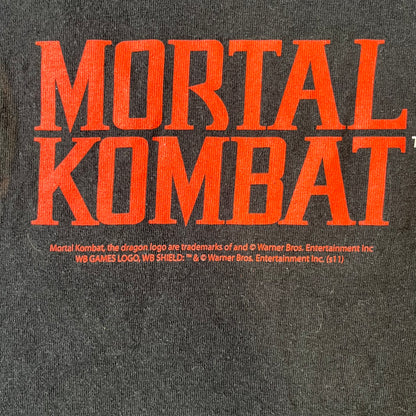 Mortal Kombat 9 Fatality Truck Tour 2011 Tee features a black T-shirt with a front graphic of a broken red ribcage and heart on a blood-covered surface. A vertical “Fatality” graphic is printed along the side, with Warner Bros. licensing text underneath the heart. One sleeve features the NetherRealm Studios logo. The back has the classic “Finish Him” text, Mortal Kombat logo, and matching licensing details.
