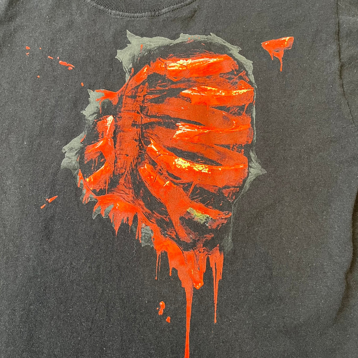 Mortal Kombat 9 Fatality Truck Tour 2011 Tee features a black T-shirt with a front graphic of a broken red ribcage and heart on a blood-covered surface. A vertical “Fatality” graphic is printed along the side, with Warner Bros. licensing text underneath the heart. One sleeve features the NetherRealm Studios logo. The back has the classic “Finish Him” text, Mortal Kombat logo, and matching licensing details.
