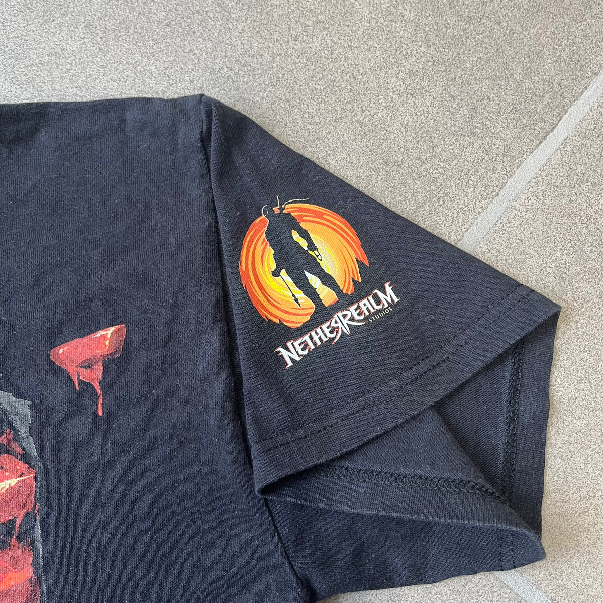 Mortal Kombat 9 Fatality Truck Tour 2011 Tee features a black T-shirt with a front graphic of a broken red ribcage and heart on a blood-covered surface. A vertical “Fatality” graphic is printed along the side, with Warner Bros. licensing text underneath the heart. One sleeve features the NetherRealm Studios logo. The back has the classic “Finish Him” text, Mortal Kombat logo, and matching licensing details.
