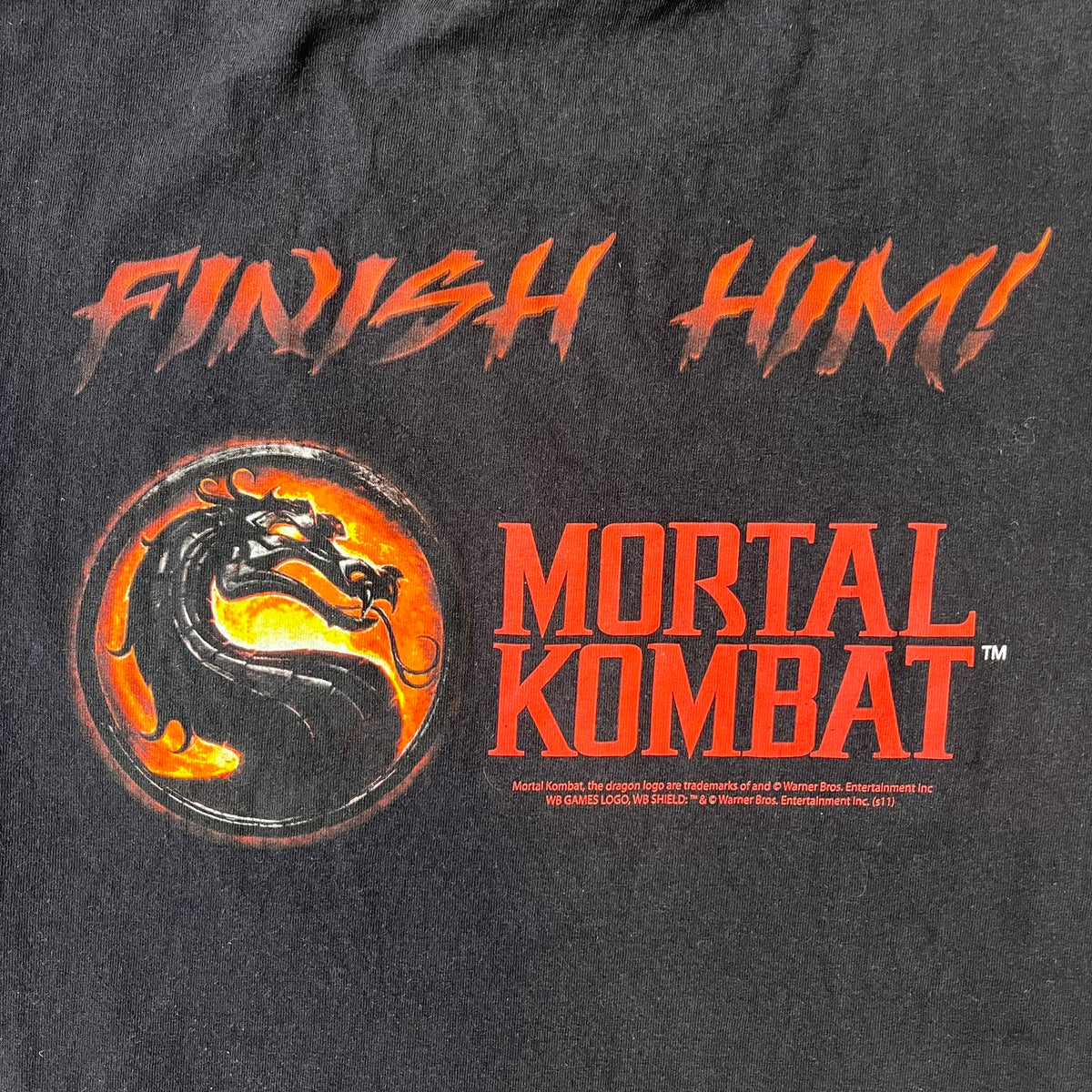 Mortal Kombat 9 Fatality Truck Tour 2011 Tee features a black T-shirt with a front graphic of a broken red ribcage and heart on a blood-covered surface. A vertical “Fatality” graphic is printed along the side, with Warner Bros. licensing text underneath the heart. One sleeve features the NetherRealm Studios logo. The back has the classic “Finish Him” text, Mortal Kombat logo, and matching licensing details.
