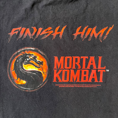 Mortal Kombat 9 Fatality Truck Tour 2011 Tee features a black T-shirt with a front graphic of a broken red ribcage and heart on a blood-covered surface. A vertical “Fatality” graphic is printed along the side, with Warner Bros. licensing text underneath the heart. One sleeve features the NetherRealm Studios logo. The back has the classic “Finish Him” text, Mortal Kombat logo, and matching licensing details.

