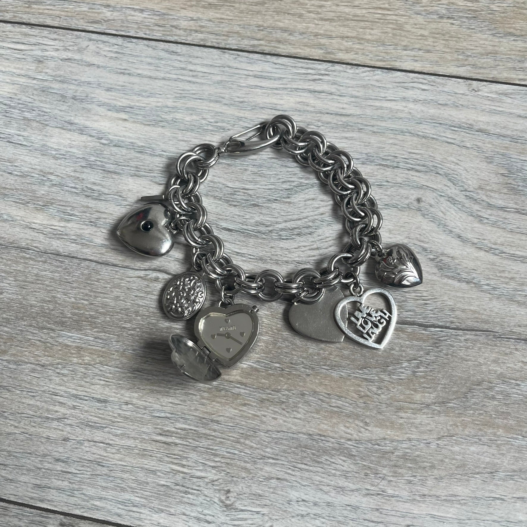 Moschino 2000s “Live Love Laugh” Charms Bracelet Watch is a stainless steel bracelet watch with a silver-tone finish. The bracelet is made of connected circular links and features multiple hanging heart-shaped charms. One charm displays “Live Love Laugh” text, while the main charm is a heart-shaped opening watch case with an “M” on the front. The watch dial is small and uses heart symbols in place of the main hour markers. All components are finished in silver tone.
